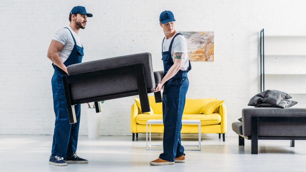 Two professional movers carefully lifting a dark gray upholstered chair in a bright, modern living room—part of a reliable Moving service in New Jersey—wearing matching navy-blue uniforms and caps, set against a clean white brick wall with a yellow sofa and minimalist decor