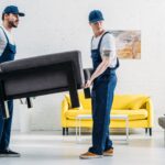 Two professional movers carefully lifting a dark gray upholstered chair in a bright, modern living room—part of a reliable Moving service in New Jersey—wearing matching navy-blue uniforms and caps, set against a clean white brick wall with a yellow sofa and minimalist decor