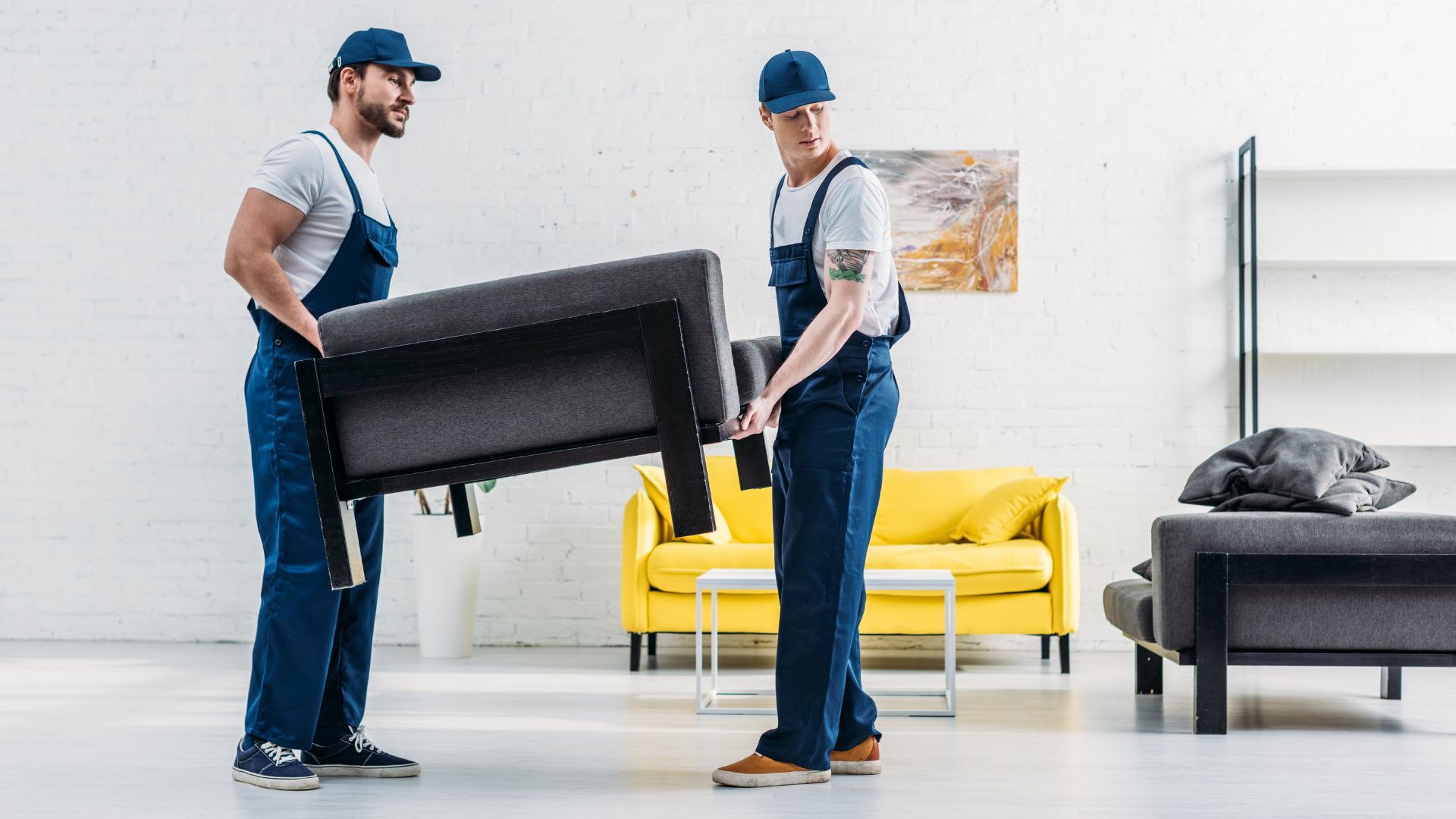Two professional movers carefully lifting a dark gray upholstered chair in a bright, modern living room—part of a reliable Moving service in New Jersey—wearing matching navy-blue uniforms and caps, set against a clean white brick wall with a yellow sofa and minimalist decor