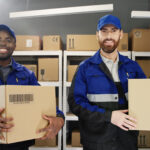 Two smiling movers Newark NJ wearing blue and black uniforms holding cardboard boxes in a warehouse filled with stacked packages on metal shelves.
