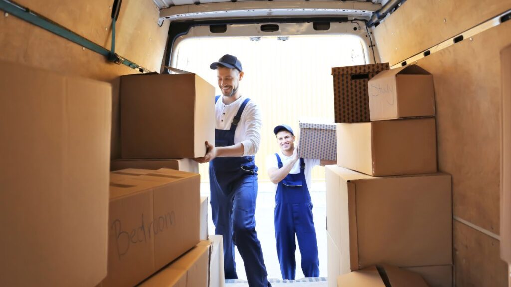Two Jersey City movers in blue overalls lift cardboard boxes into the back of a moving van. One is inside the van carrying a large box, while the other stands outside holding another box. The interior of the van is lined with wooden paneling.