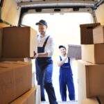 Two Jersey City movers in blue overalls lift cardboard boxes into the back of a moving van. One is inside the van carrying a large box, while the other stands outside holding another box. The interior of the van is lined with wooden paneling.