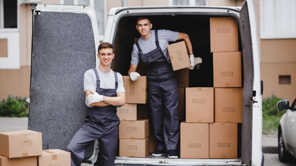 Two professional movers unloading cardboard boxes from a white moving van at a residential driveways—ideal depiction of Movers near me.