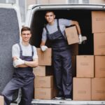 Two professional movers unloading cardboard boxes from a white moving van at a residential driveways—ideal depiction of Movers near me.