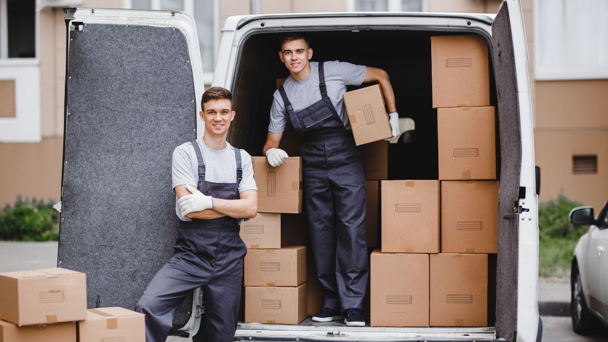 Two professional movers unloading cardboard boxes from a white moving van at a residential driveways—ideal depiction of Movers near me.