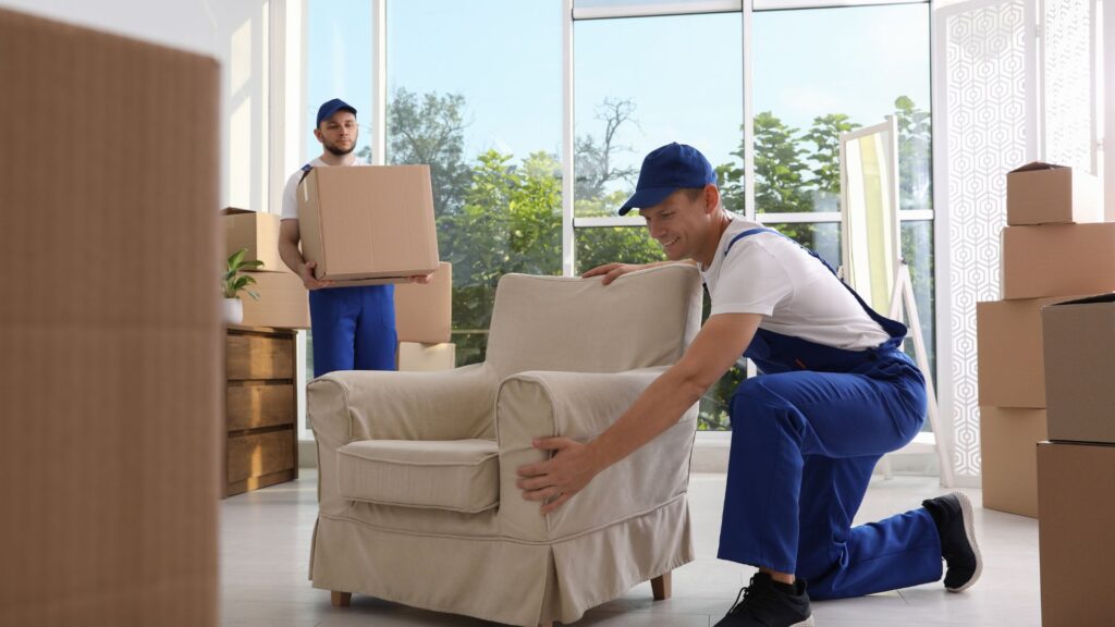 Two movers in blue uniforms carrying boxes and positioning an armchair inside a bright room during a residential move in New Jersey (movers NJ).
