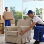 Two movers in blue uniforms carrying boxes and positioning an armchair inside a bright room during a residential move in New Jersey (movers NJ).