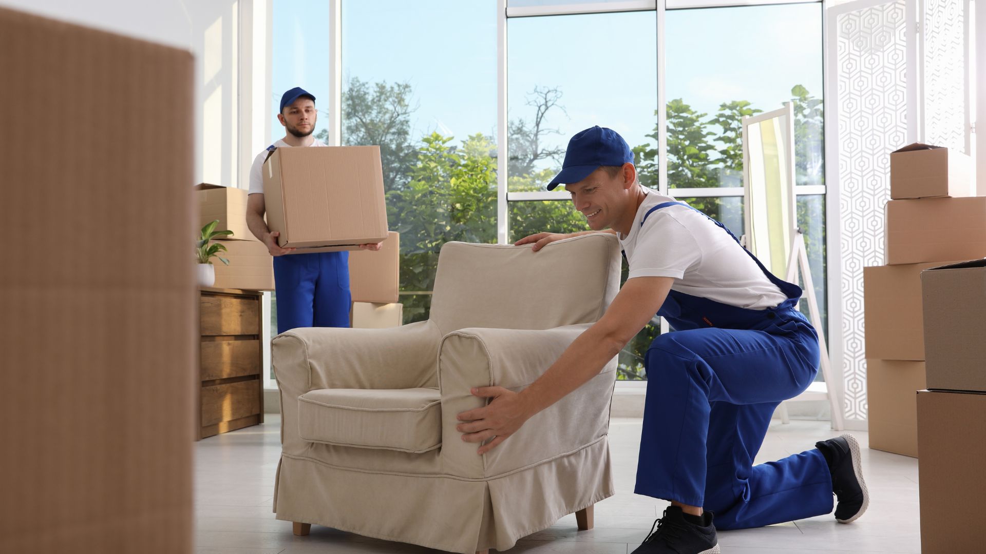 Two movers in blue uniforms carrying boxes and positioning an armchair inside a bright room during a residential move in New Jersey (movers NJ).