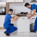 Two movers wearing blue work uniforms—Jersey City movers—carefully maneuvering a dishwasher during installation beneath a modern kitchen counter, surrounded by cabinetry and appliances.