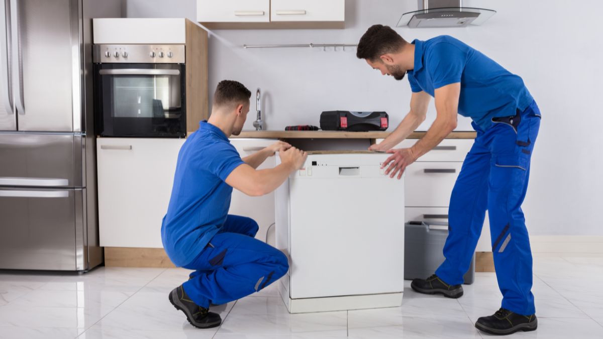 Two movers wearing blue work uniforms—Jersey City movers—carefully maneuvering a dishwasher during installation beneath a modern kitchen counter, surrounded by cabinetry and appliances.